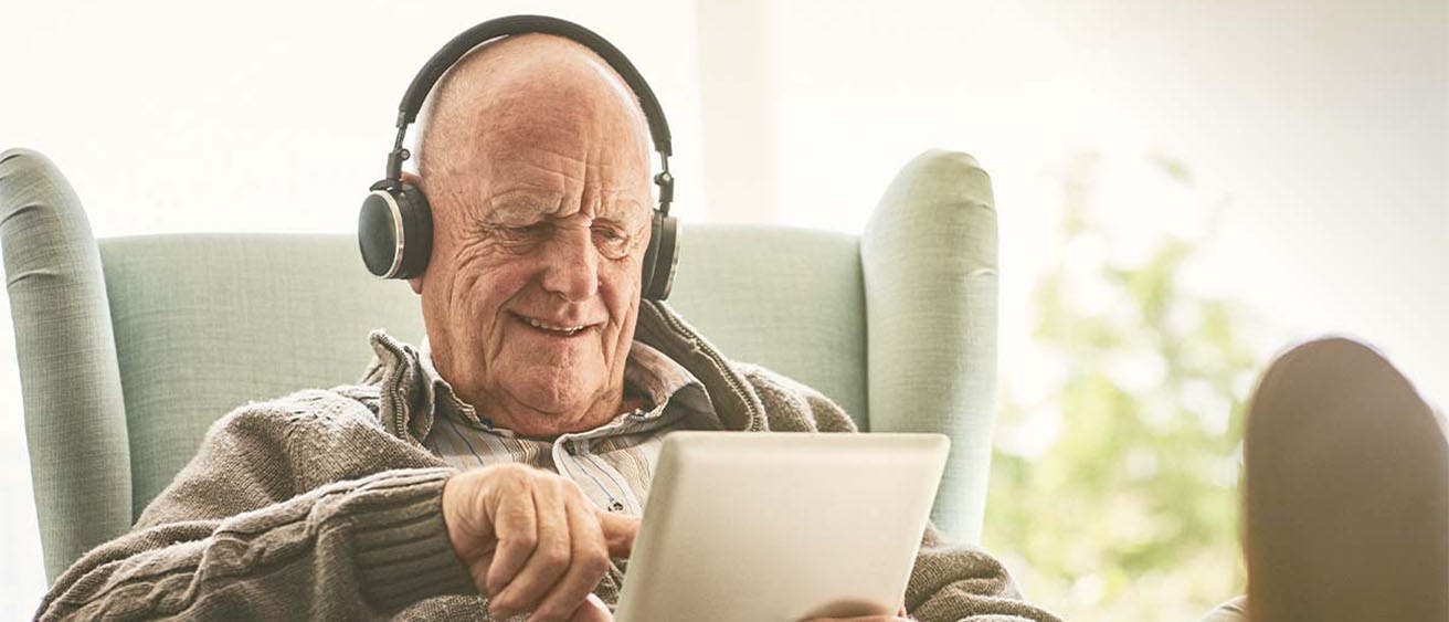 An older man wearing ear phones sitting in an armchair with a tablet.